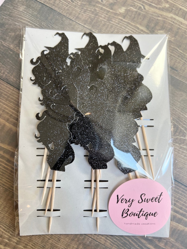 Puede incluir: Un paquete de adornos para cupcakes con silueta de bruja negra con purpurina y palitos de madera. El paquete tiene una pegatina circular rosa con el texto "Very Sweet Boutique creaciones hechas a mano".