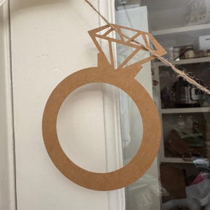 May include: A brown paper diamond ring decoration hanging from a twine string. The ring has a large circular band and a diamond-shaped top. The decoration is likely for a wedding or engagement party.