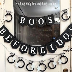 May include: Black banner with white text spelling "BOOS BEFORE I DO" and diamond ring and ghost decorations. The banner is strung across a doorway, perfect for a Halloween or spooky wedding theme.