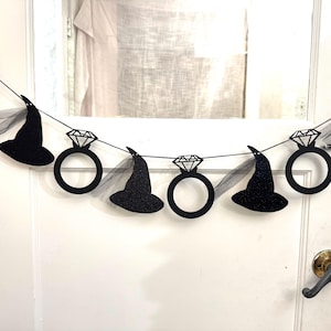May include: A black glitter banner with alternating diamond ring and witch hat cutouts. The banner is strung on a black string and includes sheer black fabric accents. The banner is hanging on a white door.