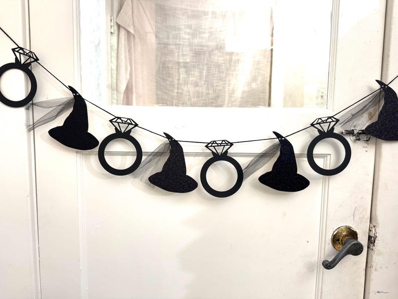 May include: A black glitter banner with alternating diamond ring and witch hat cutouts. The banner is strung on a black string and includes sheer black fabric accents. The banner is hanging on a white door.