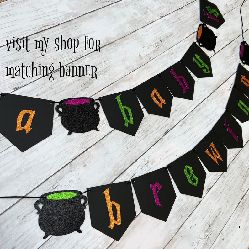 Puede incluir: Una pancarta de baby shower de Halloween negra y verde con las palabras "a baby is brewing" y decoraciones de caldero.
