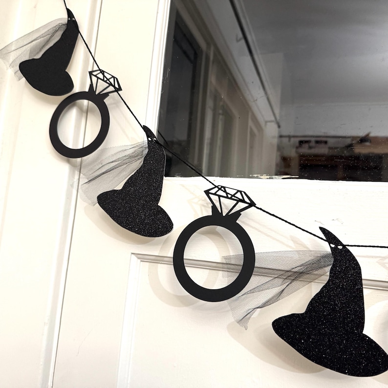 May include: A black glittery witch hat and diamond ring banner. The banner is strung on a black string and has a sheer black veil attached to the hats. The banner is hanging in front of a white door and window.