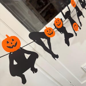 May include: A Halloween banner featuring black silhouettes of figures with orange jack-o'-lantern heads. The banner is strung on a black string and is against a white background. The jack-o'-lanterns have carved faces.