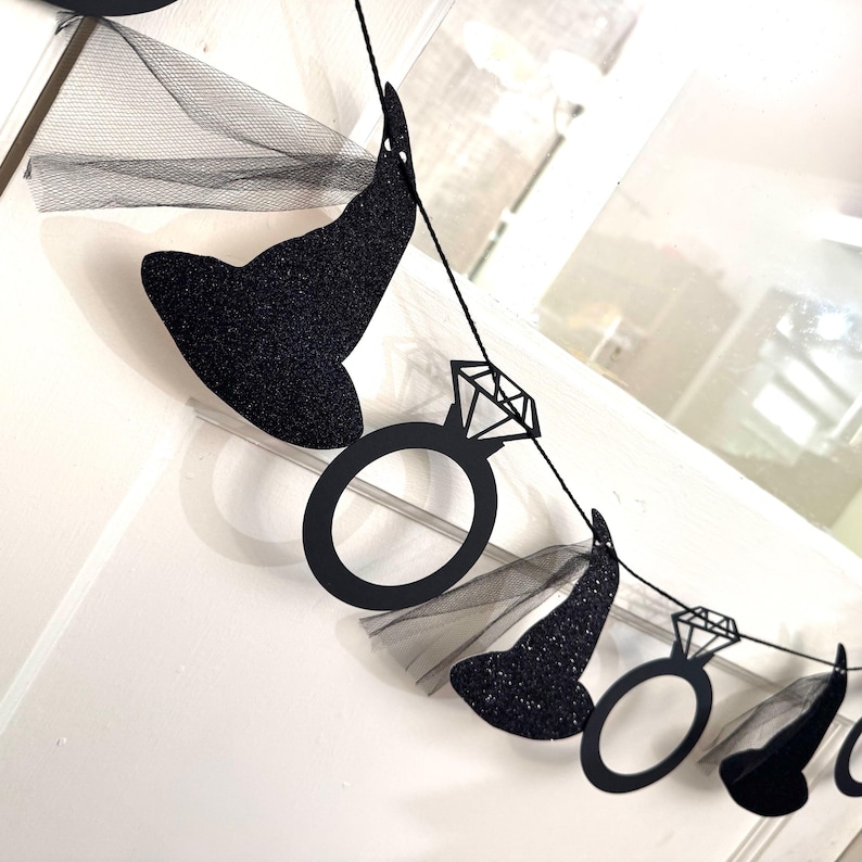 May include: A black glitter witch hat and diamond ring banner. The banner is strung on a black string and includes black tulle. The witch hats are covered in black glitter. The diamond rings are black cutouts.