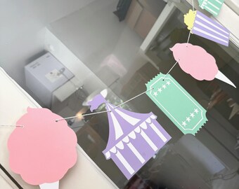 May include: A festive banner featuring circus-themed cutouts. The banner includes a pink cotton candy, a light purple and white circus tent, popcorn, and ticket stubs. The banner is strung on a white string and is perfect for a party.