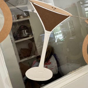 May include: A paper cocktail glass decoration with a brown drink and white stem. The decoration is strung on twine and is part of a banner. The background includes a window and a glimpse of a kitchen.