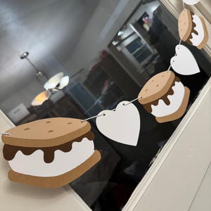 May include: A decorative banner featuring paper cutouts of s'mores and white hearts strung together with string. The s'mores have brown graham cracker layers, white marshmallow filling, and chocolate. The banner is designed for festive occasions.