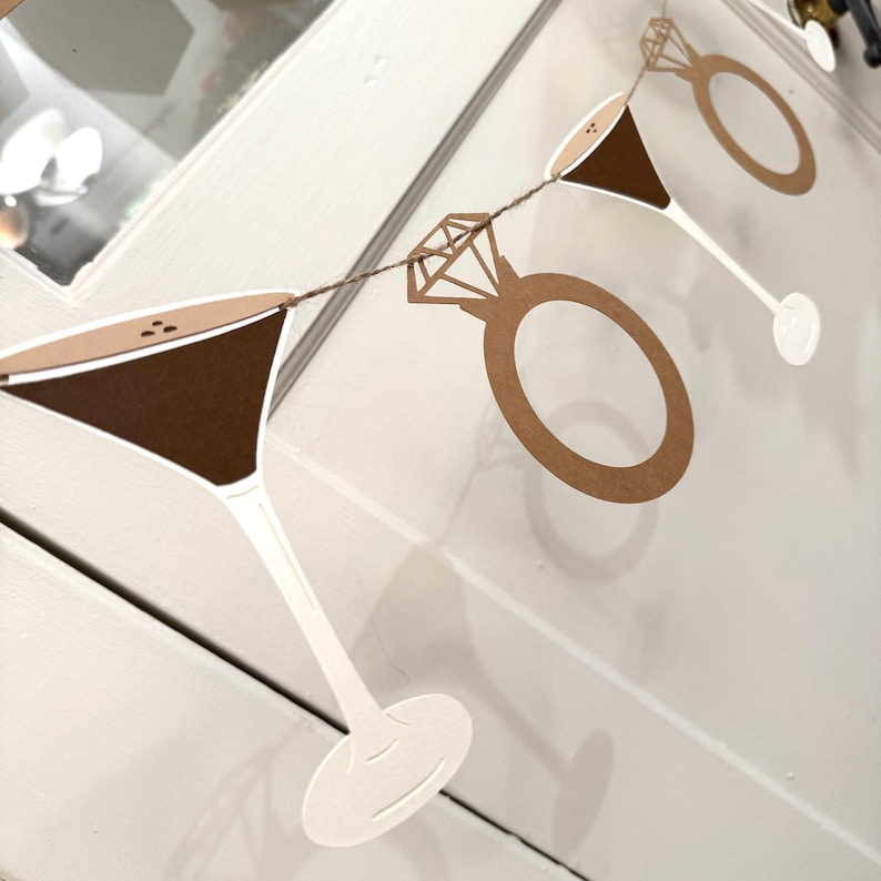 May include: A decorative garland featuring cut-out martini glasses and diamond rings. The martini glasses have brown filling and white stems. The rings are a light brown color and are connected by a brown string. The garland is perfect for a bridal shower.