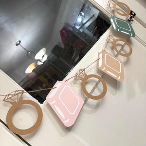 May include: A decorative banner featuring alternating diamond ring and book-shaped cutouts. The rings are a light brown color, and the books are pink, green, and tan with white borders. The banner is strung on a thin, natural-colored twine.