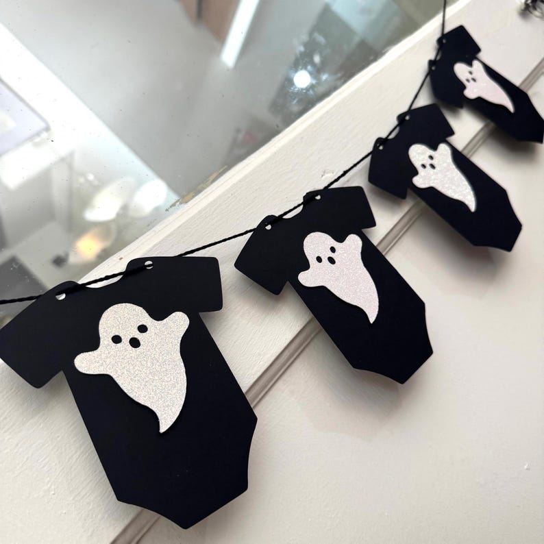 May include: A Halloween-themed banner featuring black onesie cutouts with white glitter ghost designs. The banner is strung on a black string, perfect for festive decorations. The onesies are arranged in a row, creating a spooky yet cute aesthetic.