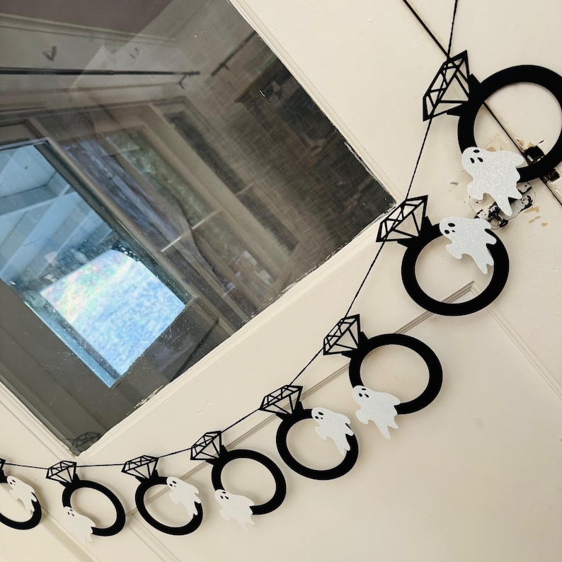 May include: A black and white Halloween banner with diamond ring and ghost cutouts. The banner is strung on black string and is hanging on a white door. The diamond rings have a geometric design.