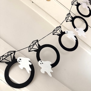 May include: A black and white Halloween banner featuring diamond ring shapes and glittery white ghost cutouts. The banner is strung on a black string and is perfect for Halloween or a spooky engagement party.