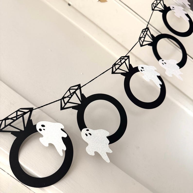 May include: A black and white Halloween banner featuring diamond ring shapes and glittery white ghost cutouts. The banner is strung on a black string and is perfect for Halloween or a spooky engagement party.