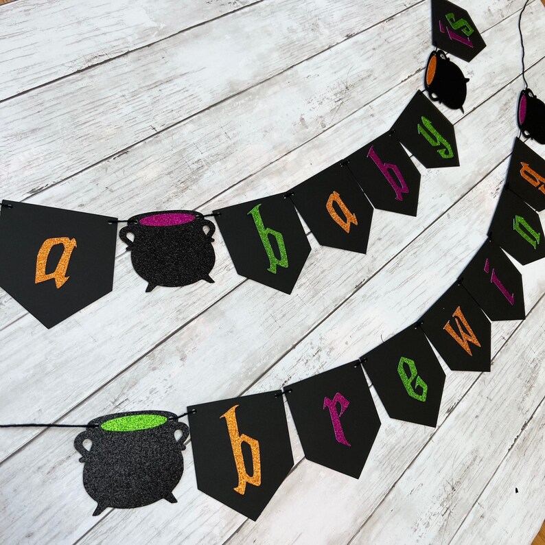 Puede incluir: Una pancarta de baby shower de Halloween negra y verde con el texto "a baby is brewing". La pancarta tiene banderines negros con letras de purpurina dorada, verde y morada. Cada bander&iacute;n tiene un caldero negro con purpurina verde o morada en su interior.
