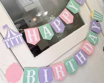 May include: A pastel-colored banner with the words "Happy Birthday" in white letters. The banner is decorated with a purple tent, a pink cotton candy, and popcorn.