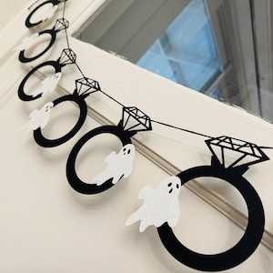 May include: A black and white Halloween banner featuring diamond ring shapes with a diamond accent and a small white ghost attached to each ring. The banner is strung on a black string and is hanging on a white door.