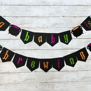 Puede incluir: Una pancarta de baby shower de Halloween negra y verde con el texto "a baby is brewing" y decoraciones de caldero negro.