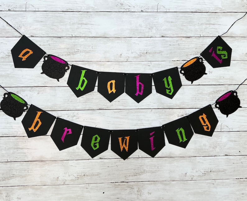 Puede incluir: Una pancarta de baby shower de Halloween negra y verde con el texto "a baby is brewing" y decoraciones de caldero negro.