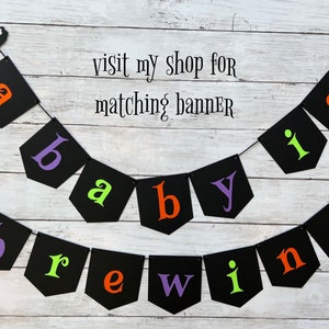 Puede incluir: Una pancarta de baby shower de Halloween en blanco y negro con el texto "a baby is brewing". La pancarta est&aacute; hecha de cartulina negra con letras blancas y tiene una silueta de bruja y un fantasma en cada extremo.