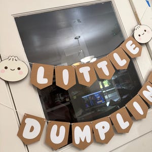 May include: A brown banner with white lettering that reads "LITTLE DUMPLING" is strung across a white door. The banner has a dumpling graphic on each end. The dumplings have smiling faces and pink cheeks.