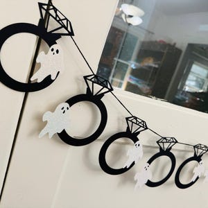 May include: A black and white Halloween banner featuring diamond ring shapes and glittery white ghost cutouts. The banner is strung together with black string and is hanging against a white background.