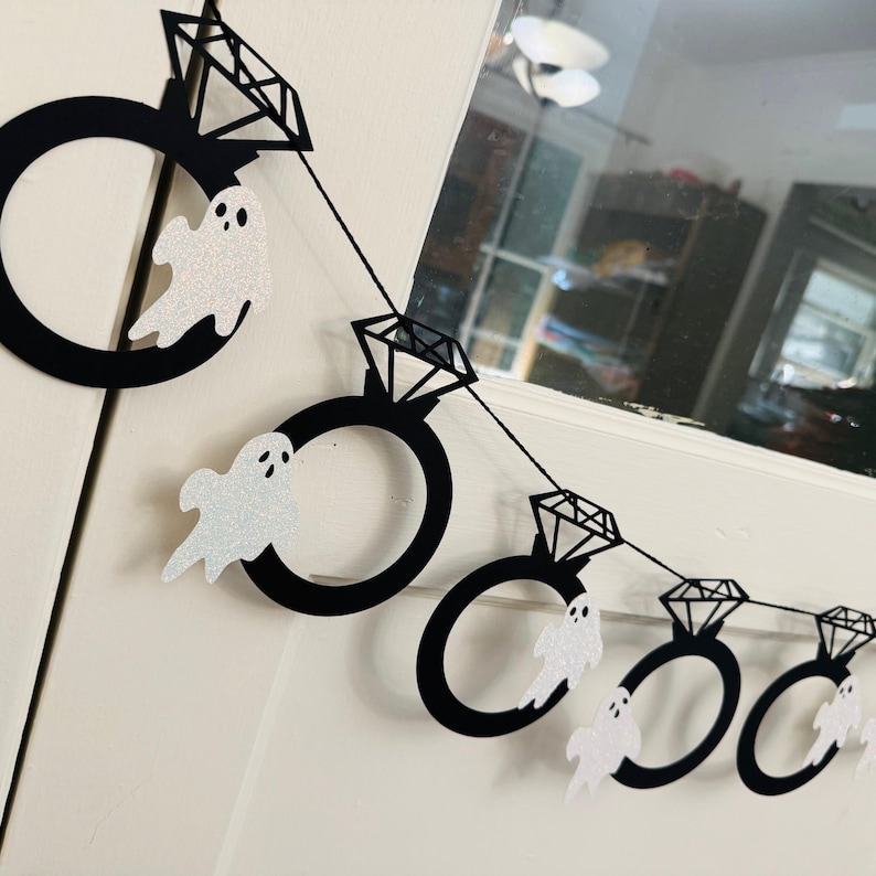 May include: A black and white Halloween banner featuring diamond ring shapes and glittery white ghost cutouts. The banner is strung together with black string and is hanging against a white background.
