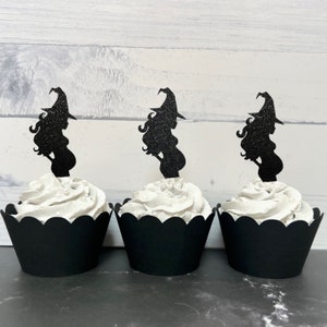 Puede incluir: Tres envoltorios de cupcake negros con glaseado blanco y adornos de cupcake de silueta de bruja negra con purpurina.