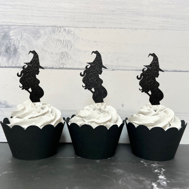 Puede incluir: Tres envoltorios de cupcake negros con glaseado blanco y adornos de cupcake de silueta de bruja negra con purpurina.