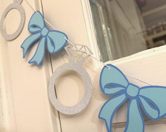 May include: A decorative banner featuring silver glitter engagement rings and light blue bows strung together with white string. The banner is designed for a wedding or engagement celebration.