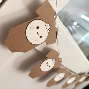 May include: A garland of brown paper cutouts shaped like baby onesies, each featuring a smiling dumpling face with pink cheeks and black eyes. The garland is strung on a thin, silver string, perfect for a baby shower or nursery decoration.
