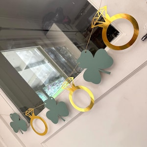 May include: A decorative garland featuring gold-colored diamond rings and green shamrock cutouts. The garland is strung on a thin, natural-colored string and is hanging on a white door. The rings have a shiny, metallic finish.