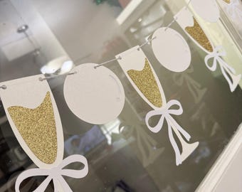May include: A white and gold glitter champagne glass banner with white bows. The banner is strung on a thin, light-colored string. The champagne glasses have gold glitter filling the glass shape. The banner is for a celebration.