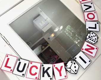 May include: A decorative banner spelling "LUCKY" and "LOVE" with playing card-shaped cutouts. The cards have red and black letters and suit symbols. Two dice are interspersed between the cards. The banner is strung across a white door.