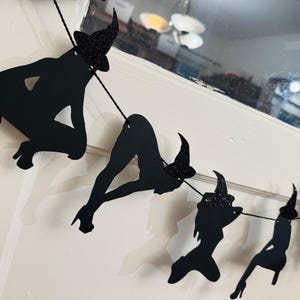 May include: Black silhouette witch garland with glittery hats. The garland features various poses of witches, strung together on a black string. Perfect for Halloween decorations.