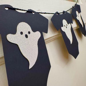 May include: A Halloween banner featuring black paper cutouts shaped like t-shirts, each adorned with a glittery white ghost. The banner is strung together with black twine, creating a festive and spooky decoration for the holiday.