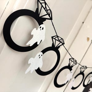 May include: A Halloween-themed banner featuring black ring shapes with diamond accents and glittery white ghost cutouts. The banner is strung together with black string, creating a festive decoration.