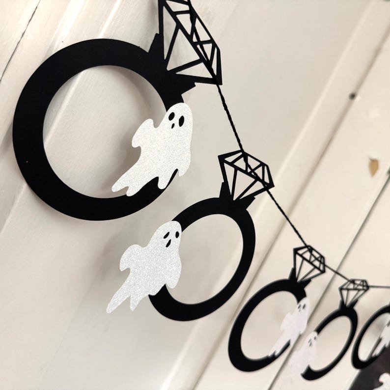 May include: A Halloween-themed banner featuring black ring shapes with diamond accents and glittery white ghost cutouts. The banner is strung together with black string, creating a festive decoration.