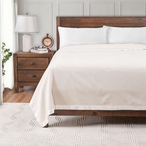 May include: A cream-colored blanket with a satin border is draped over a wooden bed frame. The bed is made with white pillows. A wooden nightstand with a lamp and a red clock is next to the bed. The rug is white with a geometric pattern.