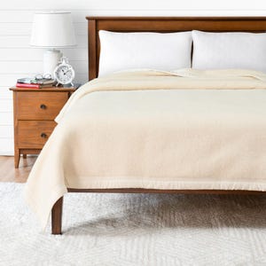 Peut inclure: Une couverture en laine de couleur crème sur un cadre de lit en bois avec des oreillers blancs. Le lit est dans une chambre avec une table de chevet en bois et une lampe blanche.