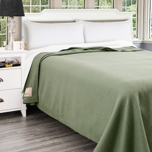 May include: A green wool blanket is folded on a white bed with two white pillows. The blanket is on a bed frame with a white headboard. A white nightstand with a lamp and a cup is next to the bed.