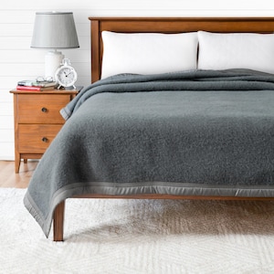Peut inclure: Une couverture grise avec une bordure grise est drapée sur un cadre de lit en bois. Deux oreillers blancs sont visibles. Une table de chevet en bois avec une lampe et un réveil est sur le côté gauche du lit. Le lit est sur un tapis blanc.