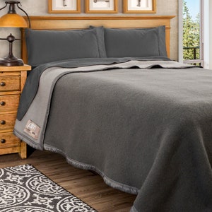May include: A gray wool blanket with a reversible design. One side is a solid gray, and the other side is a lighter gray. The blanket is folded on a bed with gray pillows.