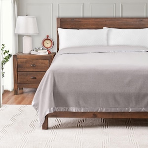 May include: A gray blanket with a satin border is draped over a wooden bed frame. The bed is made with white pillows. A wooden nightstand with a lamp and a red clock is next to the bed. The room has a neutral color scheme.