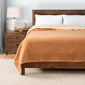 May include: A brown wool blanket with a white underside is folded on a wooden bed frame. The blanket has a fringed edge. The bed is in a bedroom with a wooden nightstand and a lamp.
