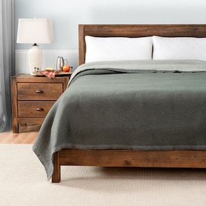 May include: A dark green wool blanket on a wooden bed frame with a white headboard. The blanket is folded back to reveal a white sheet underneath.