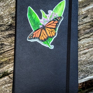 May include: A black leather-bound journal with a sticker of a monarch butterfly on a green leaf with pink flowers. The sticker says "Danaus plexippus Asclepias syriaca" and "Green Blossom Painting".
