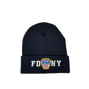 Op de afbeelding: Een zwarte gebreide muts met de witte letters "FDNY". Een kleurrijke patch met de skyline van New York City en een brandweerembleem is gecentreerd tussen de letters.