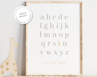 May include: A printable alphabet chart with a light gray background and a light pink 't' for 'tate'. The letters are in a simple, modern font. The chart is framed in a light wood frame.