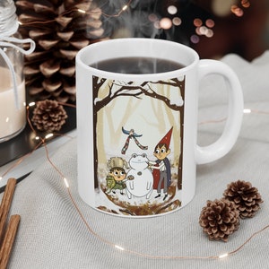 May include: White ceramic mug with a winter scene featuring two characters building a snowman in a snowy forest. The characters are wearing winter clothing and the snowman is wearing a scarf. The mug has a black handle and is filled with coffee.